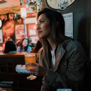 Young woman in dive bar restaurant holding beer in hand full glass watching looking drinking